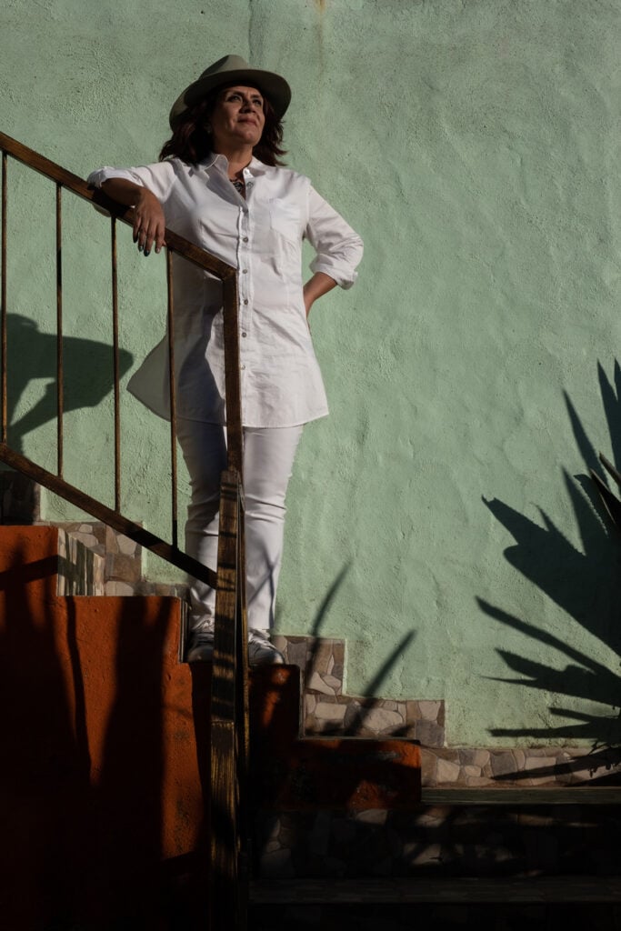 Master distiller Erika Sangeado dressed in white pants and shirt standing on orange stairs in front of a light green wall.