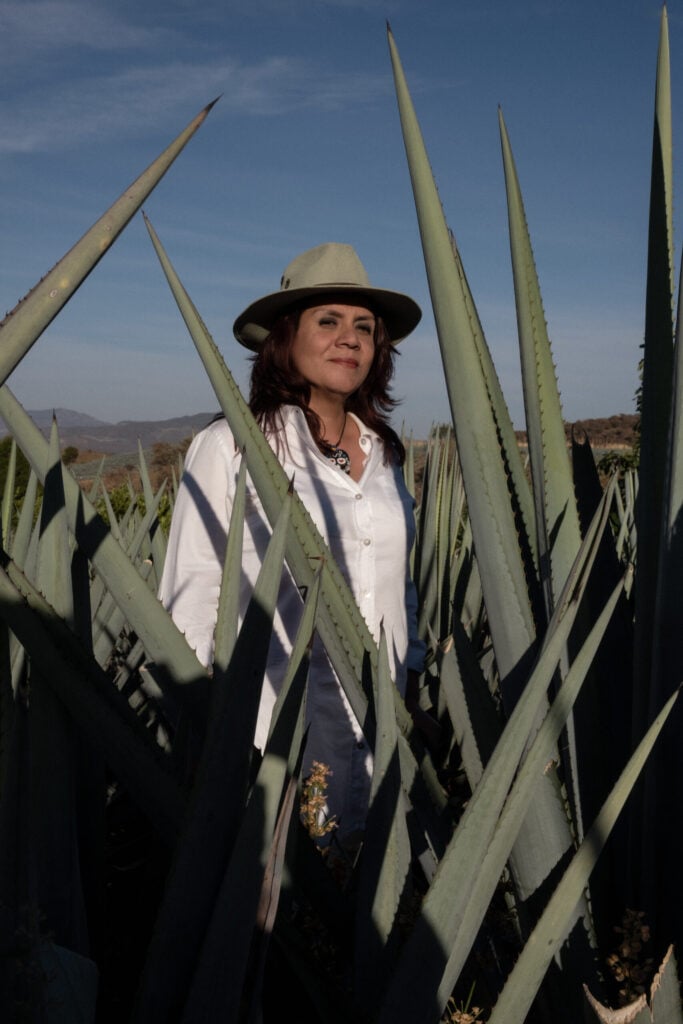 Master distiller Erika Sangeado standing in an agave field. She wears a white shirt and a light brown hat. The sky is blue and clear.