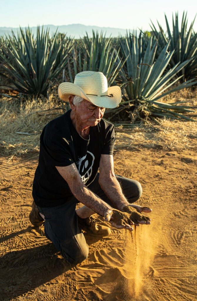 An agave farmer kneeing on the brown earth. With his two hands he holds the earth. He wears a black shirt and a beige hat.