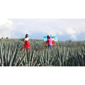 L'artista Laura Garcia Rulfo disegna Adriana in un campo di agave. Adriana indossa una gonna rossa e una camicetta bianca, mentre Laura indossa jeans blu e un maglione rosa. Sullo sfondo ci sono delle nuvole.