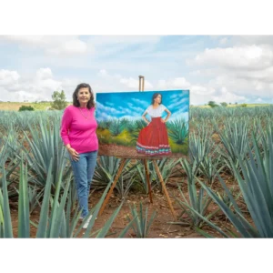 Laura Garcia Rulfo, l'artista messicana della seconda Artist Edition, in piedi in un campo di agave weber blu accanto al suo dipinto di Adriana. Laura indossa jeans blu e un maglione rosa. Il campo di agave è verde e sembra essere infinito.