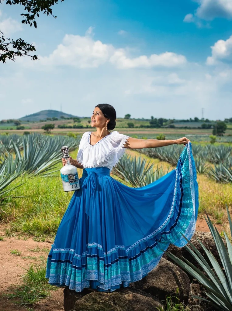 Adriana dressed in a blue skirt and white shirt holding a white Blanco bottle while standing in an agave field.